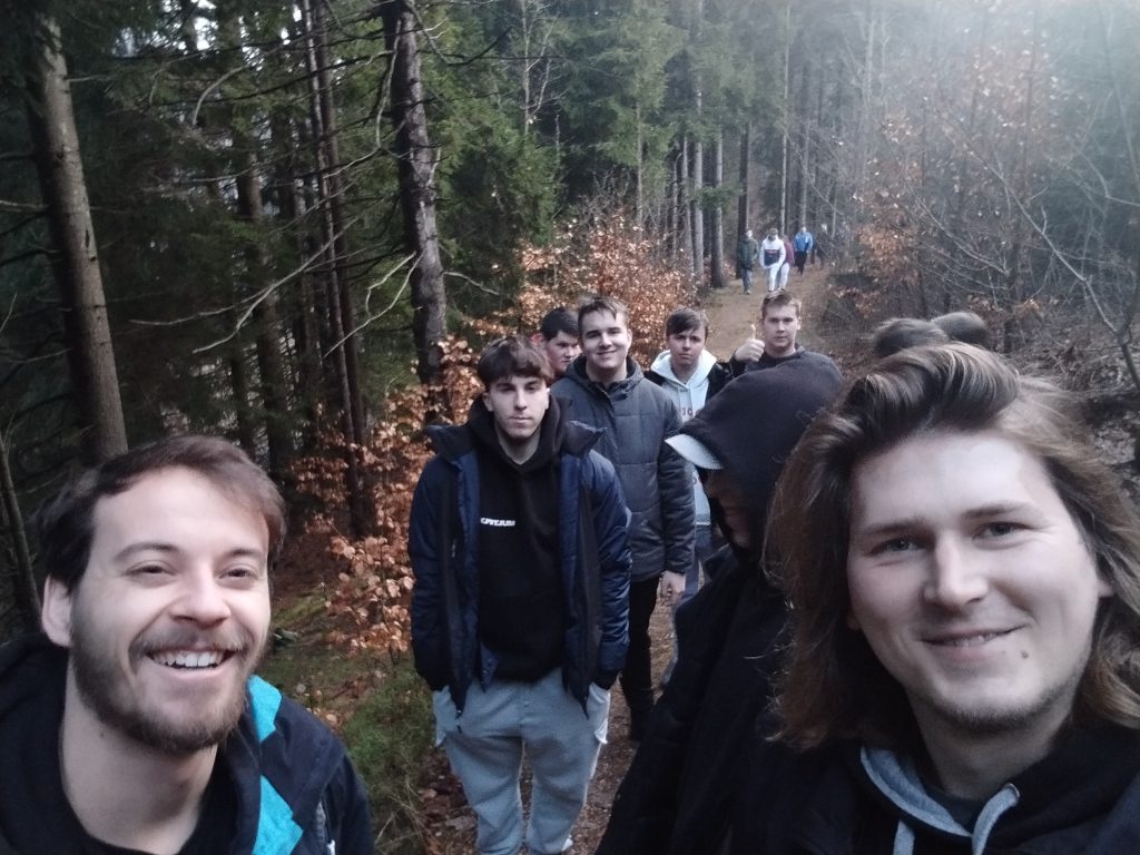 1.	Dva muži fotí selfie na lesní cestě, v pozadí na cestě jde dalších 7 osob.
