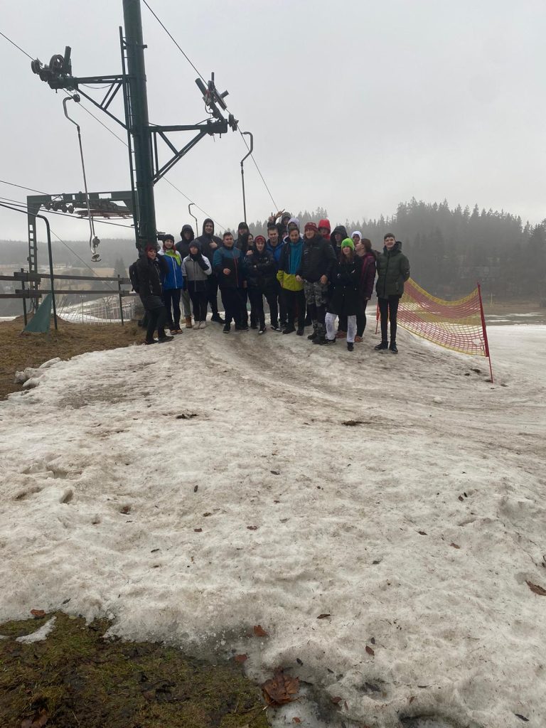 12.	Společná fotka školní skupiny 19 žáků a tří učitelů fotící se u vleku skiareálu Severák, kde byl nedostatek sněhu.