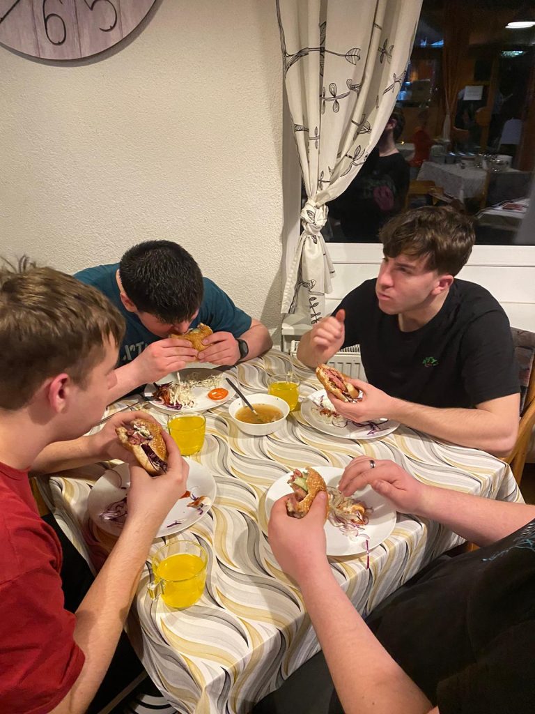 7.	Pohled na tři muže sedící v jídelně hotelu, pochutnávající si na hamburgerech.