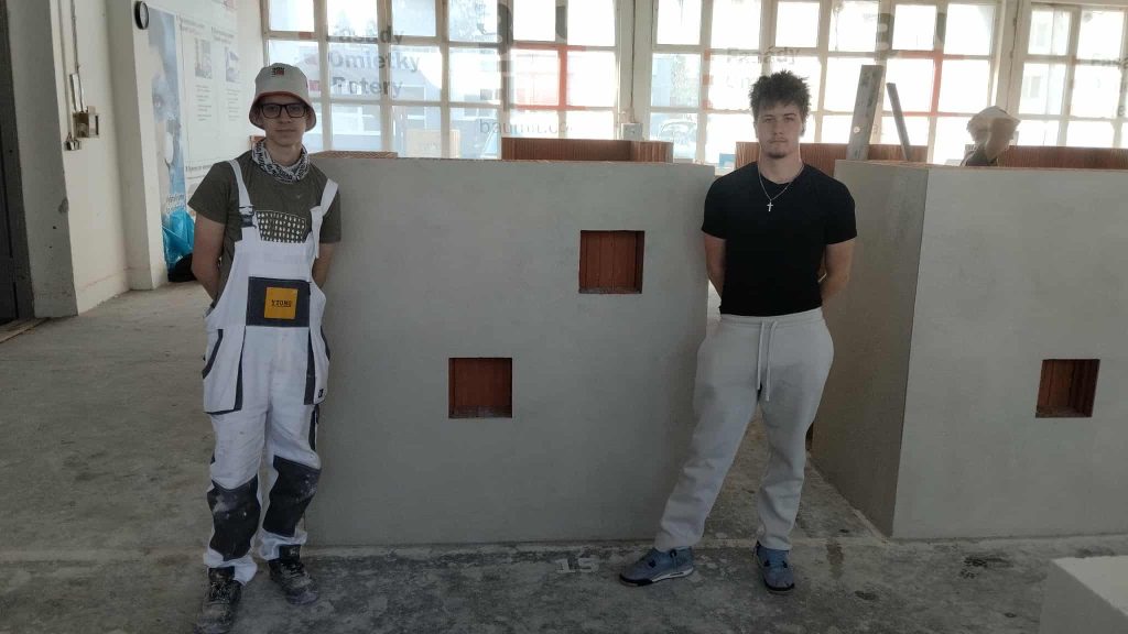 Na fotografii jsou dva žáci stojící u dokončeného soutěžního úkolu – omítnuté zdi s otvory, kterou zhotovili v rámci soutěže.